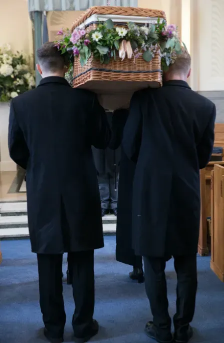 Porteurs en manteau noir portant un cercueil en osier fleuri pendant une cérémonie.